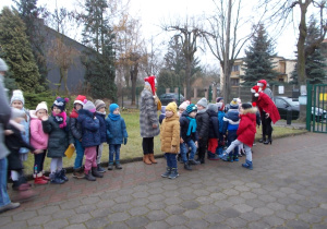 Wszystkie dzieci ubrane stoją na chodniku przed przedszkolem ustawione w szeregu.