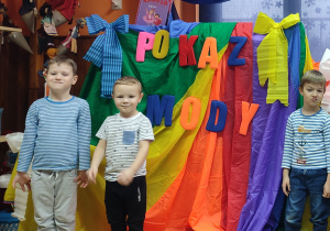 Trzech chłopców w bluzkach w paski pozuje do zdjęcia. Za nimi dekoracja: kolorowa chusta z przypiętymi literami tworzącymi napis POKAZ MODY.