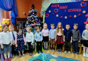 Krzesłą ustawione w półkolu. Przed nimi stoją dieci. Z tyłu dekoracja: ubrana choinka i napis na niebieskim tle: Dla Babci i Dziadka.