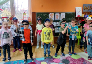 Dzieci stoją na dywanie. Ubrani w bluzy, czapki z daszkiem i okulary rapują piosenkę.