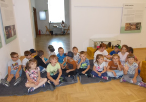 Dzieci w sali muzealnej. Siedzą na kocach.