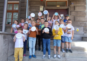 Cała grupa stoi na schodach przed budynkiem muzeum. Podnoszą do góry swoje talerzyki.