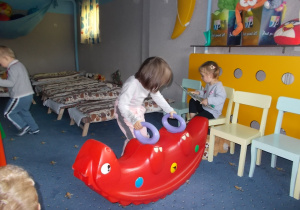 Dwie dziewczynki. Jedna siedzi na krzesełeczku i ogląda książeczkę. Druga przysuwa do niej plastikową huśtawkę.