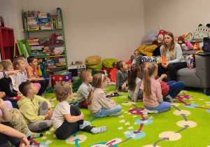 Dzieci siedzą na dywanie w jednej z sal biblioteki. Słuchają opowiadań pani będącej pracownikiem placówki.