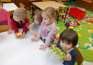 Grupka dzieci stoi przy stoliku. Jedna dziewczynka trzyma pojemnik z winogronem.