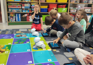 Dzieci siedzą przed matą. Każdy pracuje na swoim tablecie. Obserwują jak porusza się robot.