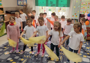 Dzieci stoją w rozsypce na dywanie. Mają białe podkoszulki. Machają żółtymi i pomarańczowymi chustami.