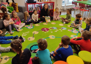 Dzieci siedzą w kole na dywanie. pani z biblioteki między nimi i pokazuje im książeczkę.