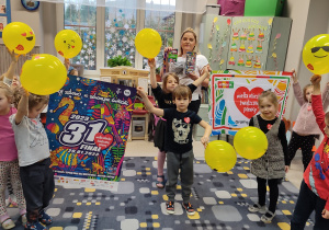 Dzieci z naklejonymi serduszkami WOŚP trzymają żółte uśmiechnięte balony.