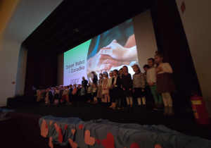 Dzieci stoją na scenie w kinie. Nad nimi obraz prezentacji multimedialnej Dzień Babci i Dziadka 2023.