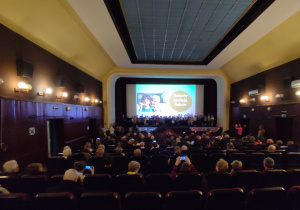 Sala kinowa sfotografowana od tyłu. Na scenie przedszkolaki a na widowni siedzą dziadkowie.