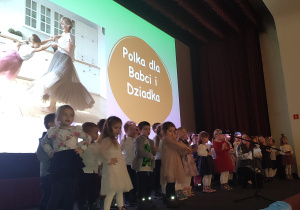 Młodsze dzieci stoją na scenie przed mikrofonem i śiewają piosenki.