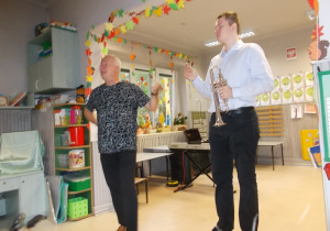 Obaj panowie stoją na środu sali. Ten z prawej strony pokazuje na instrument a ten z prawej strony pokazuje kolejne elementy budowy trąbki. W dłoni trzyma metalowy ustnik.