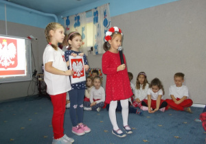 Dziewczyna w czerwonej sukience i wianku biało-czerwonym trzyma mikrofon i mówi wiersz. Za nią dwie dziewczynki trzymają tablicę z godłe.