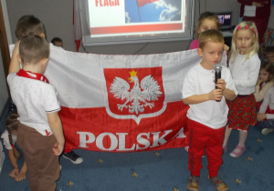 Chłopczyk trzyma mikrofon i mówi wiersz. Z tyłu dzieci trzymają rozwiniętą flagę Polski. Jeszcze bardziej z tyłu, na tablicy multimedialnej, filmik z powiewającą na wietrze flagą biało-czerwoną.