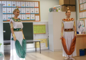 Dwie panie w białych sukniach w stylu greckim. Na głowach mają diademy a w rękach szarfę.