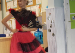 Pani w czerwono-czarnej sukience z falbanami. W ręce trzyma rozłożony czarny wachlarz.
