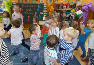 Dzieci stoją w rozsypce na dywanie w różnych pozycjach. Uśmiechają się. Na tablicy napis Noworoczny Bal z Basią. W różnych miejscach sali przymocowane duże kokardy z bibuły w paski.