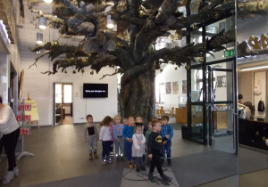 Duży hol. Na środku wysokie, metalowe drzewo. Pod nim grupka dzieci.