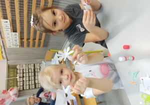 Dwie dziewczynki siedzą przy stoliku i pokazują jaki kształt gwiazdek im udało się wyciąć.