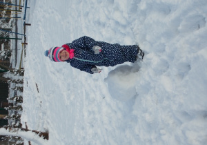 Dziewczynka w niebieskim kombinezonie w białe ciapki stoi przy śniegowej kuli. Uśmiecha się.