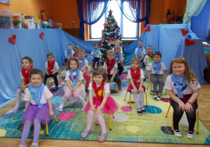 Dzieci siedzą na dywanie na krzesłach ustawionych w szachownicy. Za nimi świąteczny akcent - choinka. Każde ubrane jest w białą podkoszulkę z naklejonym serduszkiem. Akcentem zimowym są zawiązane na szyi szaliki w kolorach niebieskim i czerwonym.