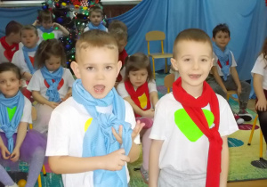 Dwóch chłopców podchodzi do kamery i recytuje wierszyk. Chłopiec z lewej strony ma szalik niebieski a chłopiec z prawej strony czerwony.