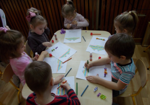 Szóstka dzieci siedzi przy stoliku. Kredkami świecowymi (niebieską, pomarańczową, zieloną) kolorują, próbując zachować rytm, paski na odrysowanej sukience.