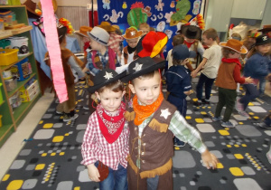Dwóch chłopców w strojach kowboja trzyma się za ramiona. Na piersi mają gwiazdy szeryfa. Takie same gwiazdy zdobią kapelusze, które mają na głowach. Uśmiechają się.