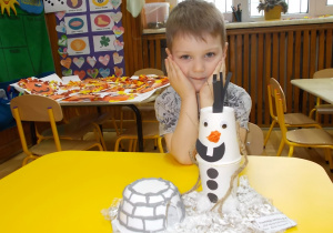 Chłopiec siedzi przy stoliku. Dłońmi podpiera głowę. Przed nim wykonana praca czyli iglo zrobione z plastikowej miski oraz bałwanek Olaf z papierowych kubków.