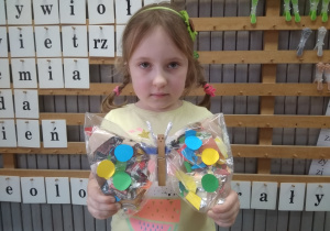Dziewczynka pokazuje motylka wykonanego z folii w środku wypełnionej kolorowymi kawałkami bibuły.