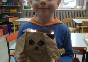 Chłopiec pokazuje pieska wykonanego z pudełka po kawie owiniętego papierem pakowym z dorysowanymi i doklejonymi elementami: uszy, łapy, oczy, wąsy, ogon.
