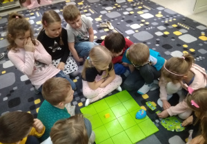 Dzieci siedzą przy małej zielonej macie. Obserwują jak porusza się po kolejnych polach robot - mała niebieska myszka.