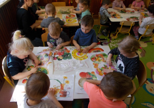 Dzieci siedzą przy stolikach na swoich miejscach. Każde ma kolorową podkładkę. W jednej ręce trzymają kawałek bursztynu, w drugiej pasek papieru ściernego. Szlifują kamień wg objaśnień.