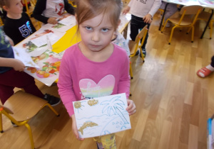Dziewczynka pokazuje swój rysunek. Słonko, chmurka i delfin pokryte są pyłem bursztynowym. Pozostałe elementy czyli woda i palma zostaną pomalowane kredkami.
