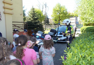 Dzieci zebrane przed przedszkolem. Na chodniku motocykl i wóz policyjny.