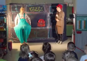 Przed sceną dwie postaci: dziewczynka ubrana w zielone spodnie z dość dużymi okrągłymi biodrami. Obok osoba w brązowym płaszczu i kapeluszu z czerwonej wełny. To babcia.