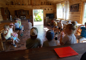 Dzieci siedzą na ławkach w drewnianym domku i czekają na spotkanie z właścicielem.