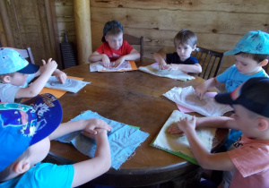 Dzieci siedzą przy stolikach. Każdy ma karton a na nim odciśniętą masę papierową, którą dodatkowo obsusza ściereczką.