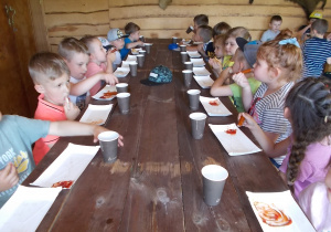 Dzieci siedzą przy długim drewnianym stole i zajadają kiełbaski z grilla.