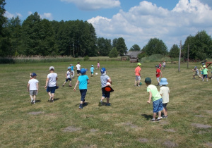 Dzieci biegają po skoszonym boisku. Jedni grają w piłkę inni ustawiają pachołki do slalomu.
