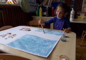 Dziewczynka siedzi przy stoliku. Przed nią duży biały karton podzielony na cztery części. Niebieską farbą maluje różne wzory.