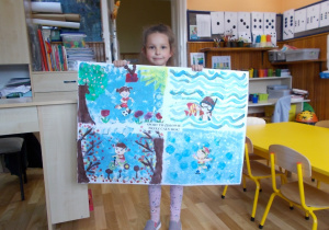 Dziewczynka pokazuje wykonaną pracę. Na niej namalowane różne dyscypliny sportowe, które można wykonywać w różnych porach roku: gra w piłkę, kąpiel w morzu, jazda na rowerze i jazda na łyżwach.