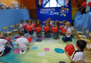 Cztery dziewczynki klęczą na dywanie. Trzymają przed sobą balony w kształcie serduszek. Chłopcy klęczą dookoła nich.