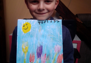 Chłopiec trzymający pracę na konkurs. Obrazek odciśniętego w farbie widelca przedstawiający pierwsze wiosenne kwiaty.