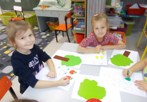 Dziewczynki przy stoliku. Kolorują pastelami wybrane owoce na swoje drzewa.