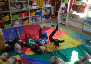 Dzieci leżą na kolorowej chuście. Wykonują nogami ćwiczenie "rowerek".