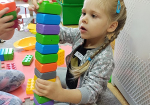 Dziewczynka w warkoczykach siedzi na dywanie i układa wieżę z kolorowych, kwadratowych klocków.