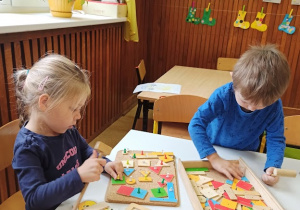 Dziewczynka i chłopczyk siedzą przy stoliku i bawią się zestawem figur geometrycznych. Przybijają elementy do korkowej tabliczki.