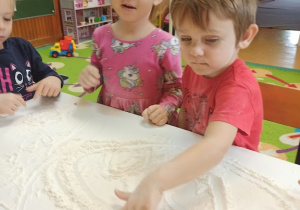 Mąka wysypana na stolnicy. Dzieci piszą po niej paluszkami tworząc różne wzory.